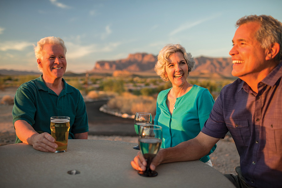 Montesa at Gold Canyon, a 55 plus community has an older woman and two older men smiling and laughing enjoying a cocktail around a table at Sunset. Beautiful Superstition mountains in background