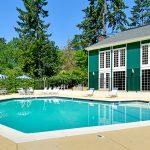 Heritage Village, an all age manufactured home community, has swimming pool with lounge chairs, tables and umbrellas. Tall lush trees all around. Green clubhouse with white shutters next to pool.