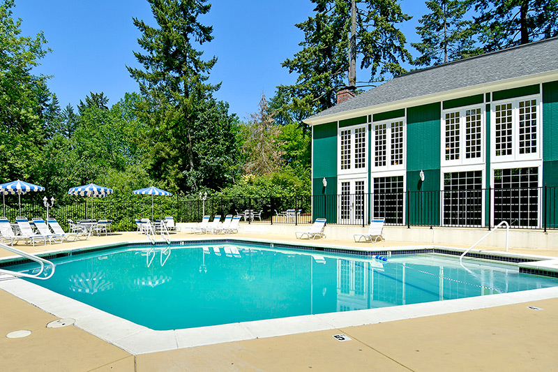 Heritage Village, an all age manufactured home community, has swimming pool with lounge chairs, tables and umbrellas. Tall lush trees all around. Green clubhouse with white shutters next to pool.
