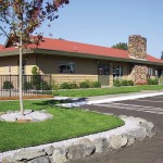 Well-maintained exterior of the community center. Trimmed trees and clean cut grass separated by stones from the paved parking lot.