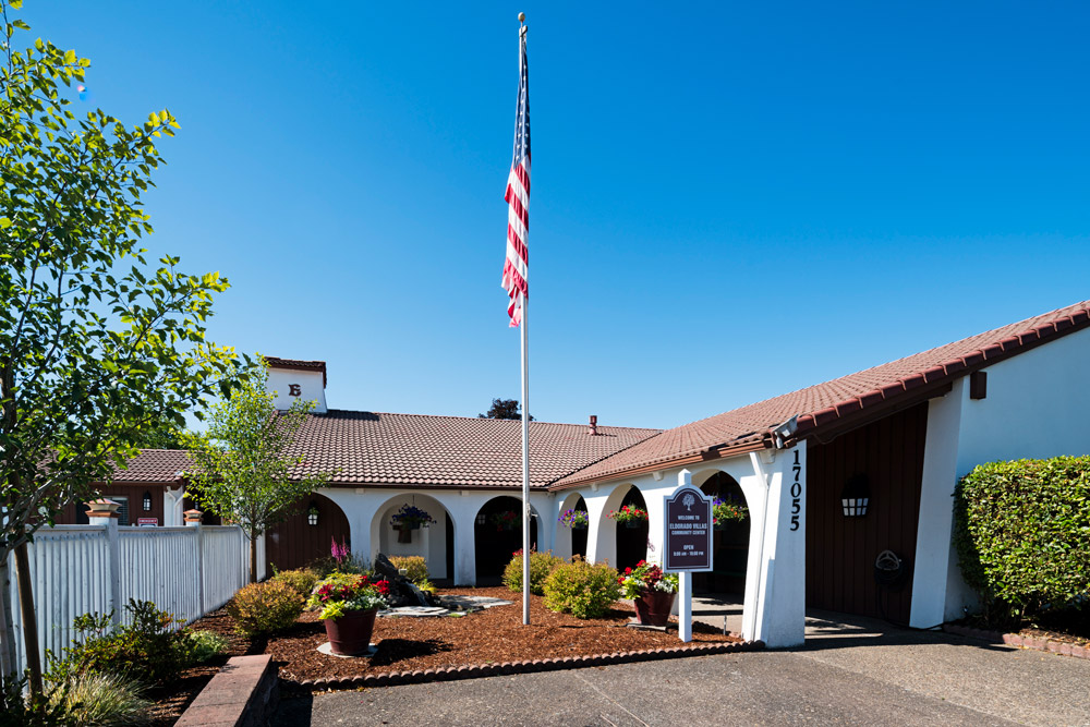 Eldorado Villas Community Center is beautifully landscape with hanging flowers of red and purple and potted plants with mixed flowers. Wood chips and small shrubs. Tall flag pole with American flag. White building with Spanish tile and archways throughout.