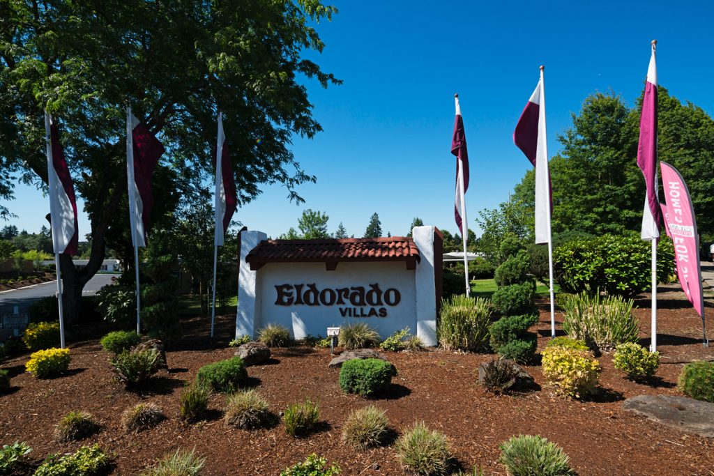 Eldorado Villas, a 55 plus manufactured home community entrance is marked with purple and white flags, small shrubs, and white sign with Eldorado Villas name. Green trees in the background.