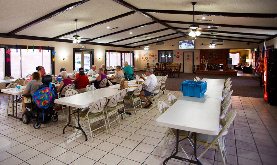 A group of older residents sit at long tables with chairs inside the clubhouse and eat lunch while creating some arts and crafts. A big screen TV mounted to the wall is on while they work.