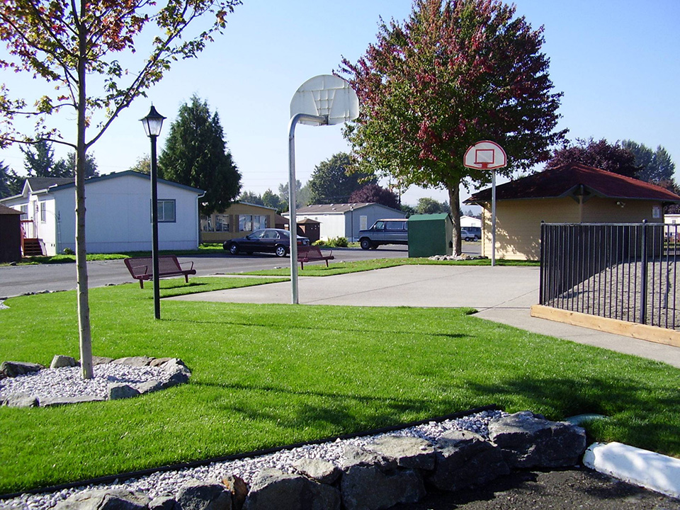 Outdoor basketball court adjacent to the playground. Surrounded by green grass and a walkway that cuts through. Two benches sit on the outer end.