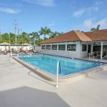 Calm swimming pool with lounge chairs and patio furniture with chairs, tables, and umbrellas. located outside clubhouse.