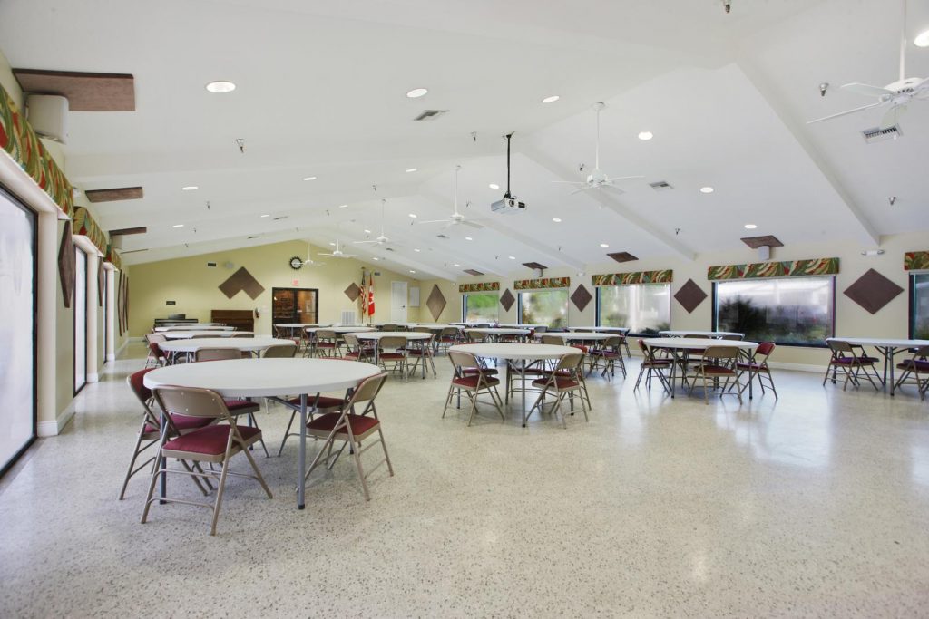 Large clubhouse with vaulted ceilings and ceiling fans. Round table with chairs cover the floor. Lots of windows on both sides.