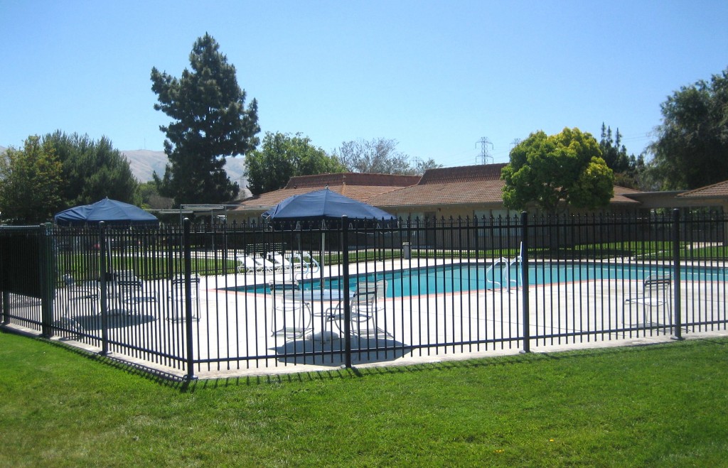 Outdoor, full-sized community pool enclosed by a black fence. Surrounded by lounge chairs and shaded tables with additional seating. Open for residents to enjoy with family and friends under the sun.