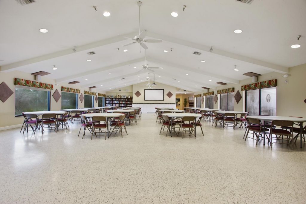 At one end of the clubhouse has large screen mounted to wall. A projector hangs from ceiling. Round tables with chairs are set up along the floor.