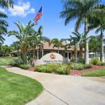 Island in the Sun, an active 55+ community has a well landscaped entrance with palm trees and green grass. An American flag flies high above the Island in the Sun sign.