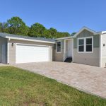 New homes are for sale at Lamplighter Village, an active 55 plus manufactured home community. Front of home with attached 2 car garage. Long driveways of pavers. Green grass and small plants for front landscape. Lush green trees in the background