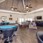 Large game area inside clubhouse with hardwood floors. Card tables with black leather chairs around them. Tall vaulted wood beamed ceilings and 4 ceiling fans. L shaped couch with 2 wood end tables and coffee table. Two decorative pillars on each side of game room. Black leather recliner chairs in front of flat screen TV mounted to wall.