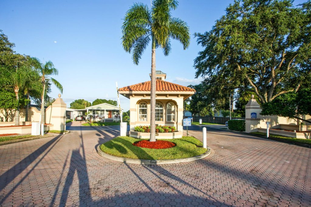 Gated entrance into Fountainview Estates, a 55 plus community. Security booth at entrance. Nicely landscaped with green grass, palm trees, full trees and brick pavers.