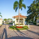 Gated entrance into Fountainview Estates, a 55 plus community. Security booth at entrance. Nicely landscaped with green grass, palm trees, full trees and brick pavers.