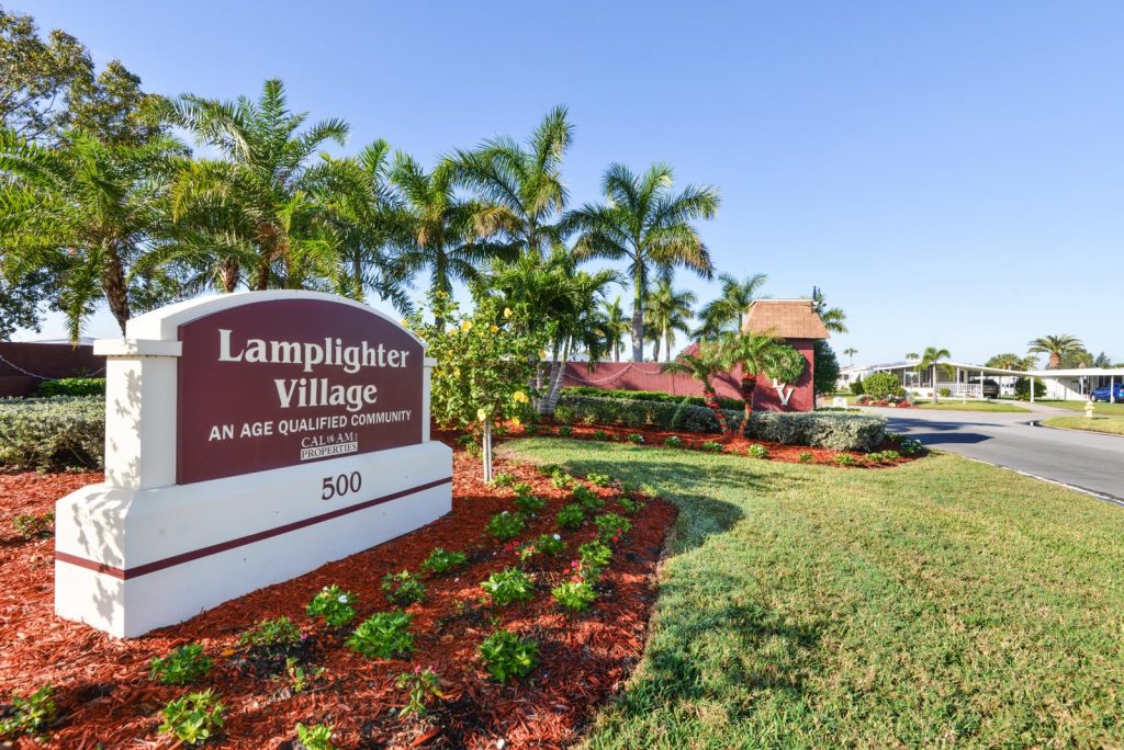 Beautifully landscaped front entrance to Lamplighter Village. Tall palm trees, green grass, and small plants surround the maroon and white stone sign indicating Lamplighter Village, an age qualified community.