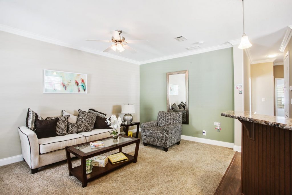 Brand new living room with light gray wall and a sage green wall. Crown molding atop and bottom. Open floor plan to the kitchen. Tan carpeting. Loveseat with white and black throw pillows and black and white ottoman chair. Dark wood coffee table and end table. Ceiling fan above coffee table.