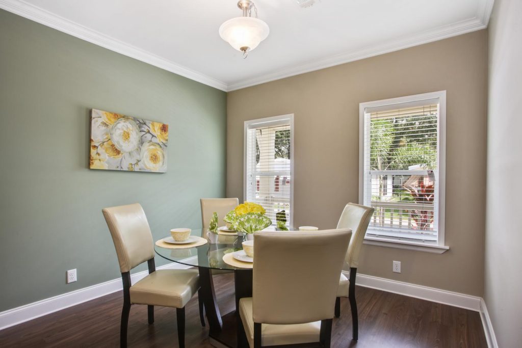 Spacious dining room that is bright and airy with 2 windows. hardwood floors with crown molding atop and bottom of walls. One wall is dark khaki in color and the other is sage green in color. Dining table is built of dark wood with circular glass top. Seating for 4 with cream, cushioned chairs