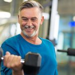 A 55+ man works out in fitness center and curls arm with free weight.