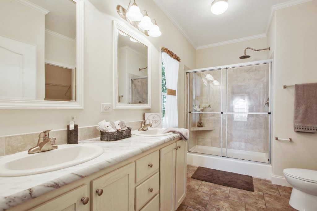 A modernized bathroom with his and her sinks. Tiled floor. Large step in shower with built-in shelf. Large showerhead. Towels are light brown with a brown shower rug.