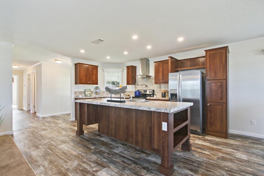 Beautiful modern kitchen with stainless steel refrigerator, range hood and oven and wood flooring throughout. Large kitchen island in the middle with granite counter tops and dark wood base. Shelves for storage and sink built into island.