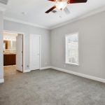 Bedroom with attached bathroom. Light gray walls with crown molding top and bottom and plush gray carpet. Two windows with blinds. Ceiling fan included.