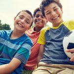 3 young boys smiling and sitting on the grass with soccer ball.