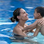 Beautiful young mom holding with excitement her son and holding him in the pool both smiling.