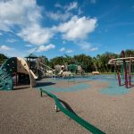 A big playground for children. There are monkey bars, slides, balance bar and swings. Bench seating is all around the perimeter of the playground. Green grass and lush trees.