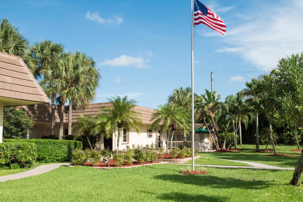 Beautiful landscape surrounds the Palm Beach Plantation. Green grass, palm trees, and lush, tall trees. Flagpole with American flag flying high in front of cluchouse.