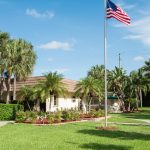 Beautiful landscape surrounds the Palm Beach Plantation. Green grass, palm trees, and lush, tall trees. Flagpole with American flag flying high in front of cluchouse.