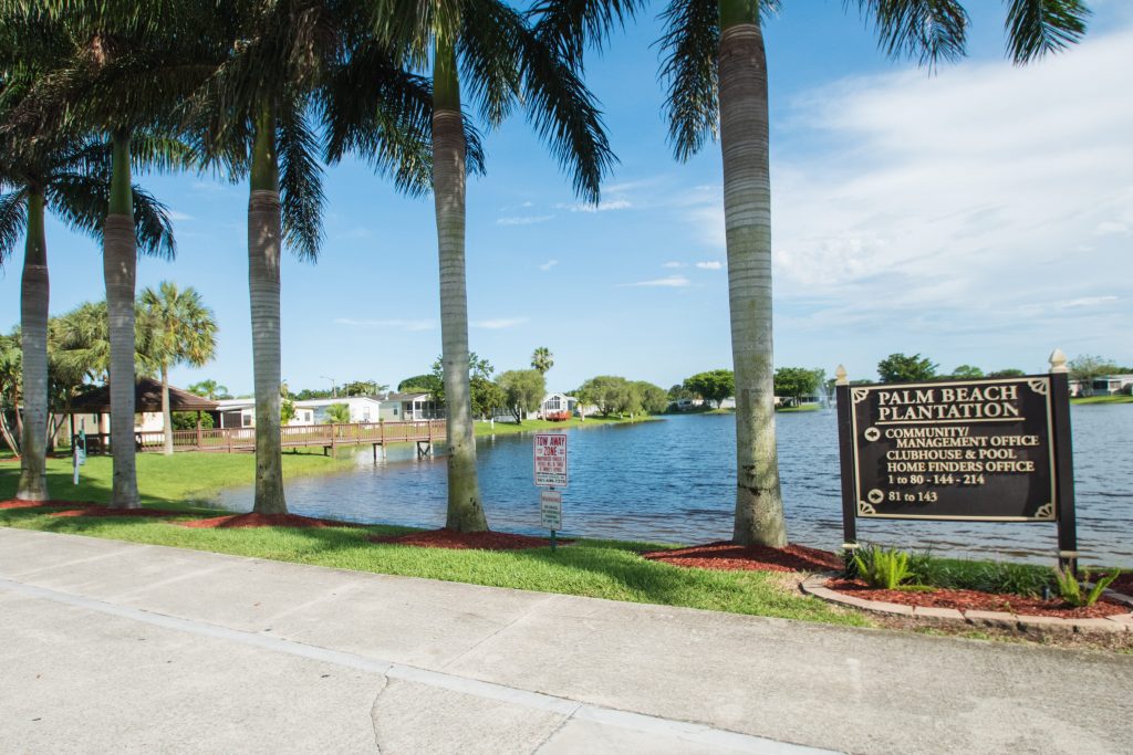 Quiet, calm waters on the lake that lap up to 5 very tall palm trees. Directional sign in front with clubhouse to the left and homes to the right well.