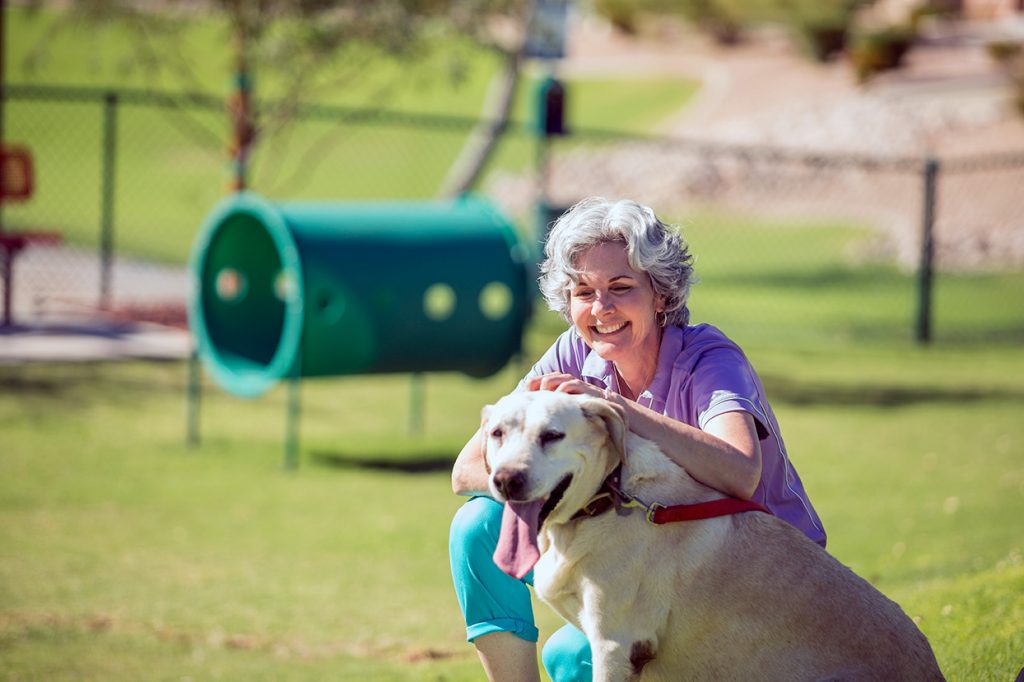 A 55 plus woman smiles big at the grassy dog park while kneeling and scratching the head of her large Labrador. Dog's tongue hangs out.