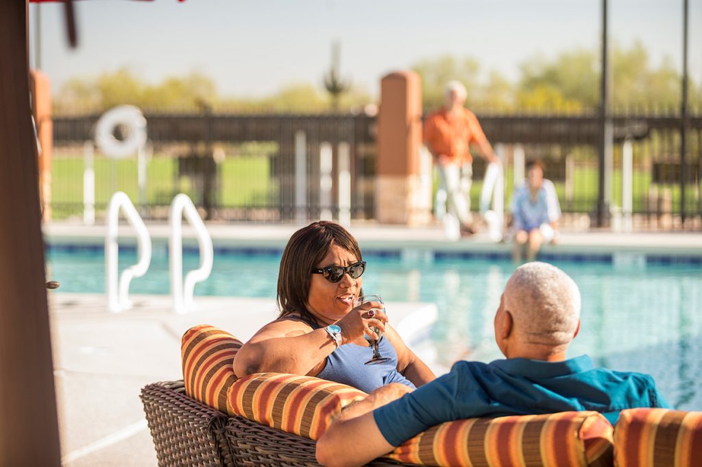 An african american couple sit in lounge seat by the pool. Woman is having a drink.