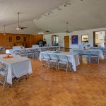 Ballroom set up with banquet tables and chairs. Vaulted ceiling with fans. White tablecloth.
