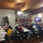 Community holiday party hosted in the community hall. 55 plus aged residents sit and enjoy dinner meals while mingling with one another. Community hall is decorated for the holidays with a Christmas tree and red and green decor over the fireplace ledge.