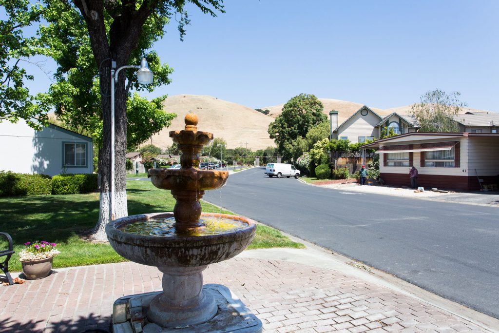 Wide, paved streets create an open space in the 55 plus community. In the foreground a water fountain is surrounded by grass and brick with the beautiful rolling hills behind.