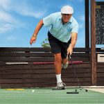 An older, retired man playing shuffleboard in black shorts, light blue polo shirt, white hat, white socks and sneakers.