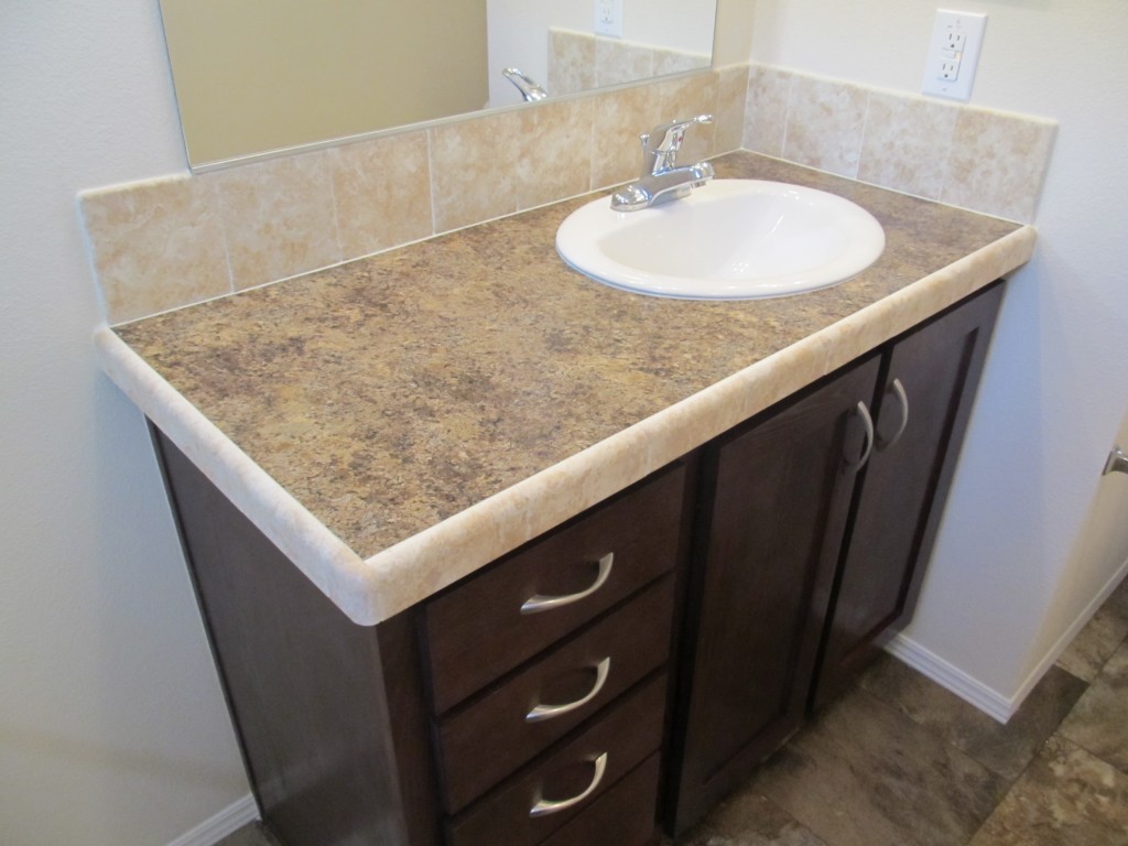 White bathroom sink surrounded by cream granite countertops, cream tile backsplash and dark walnut cabinets beneath.