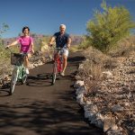 Man and woman riding bicycles along smooth path landscaped w shrubs and rocks