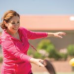 Woman in magenta sweatshirt smiling while mid swing on pickleball court.