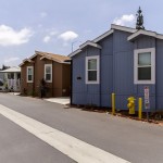 Clean and well maintained manufactured homes line the wide streets. Sky blue colored home sits in the foreground neighboring a tan home and a cream corner home with a front patio.