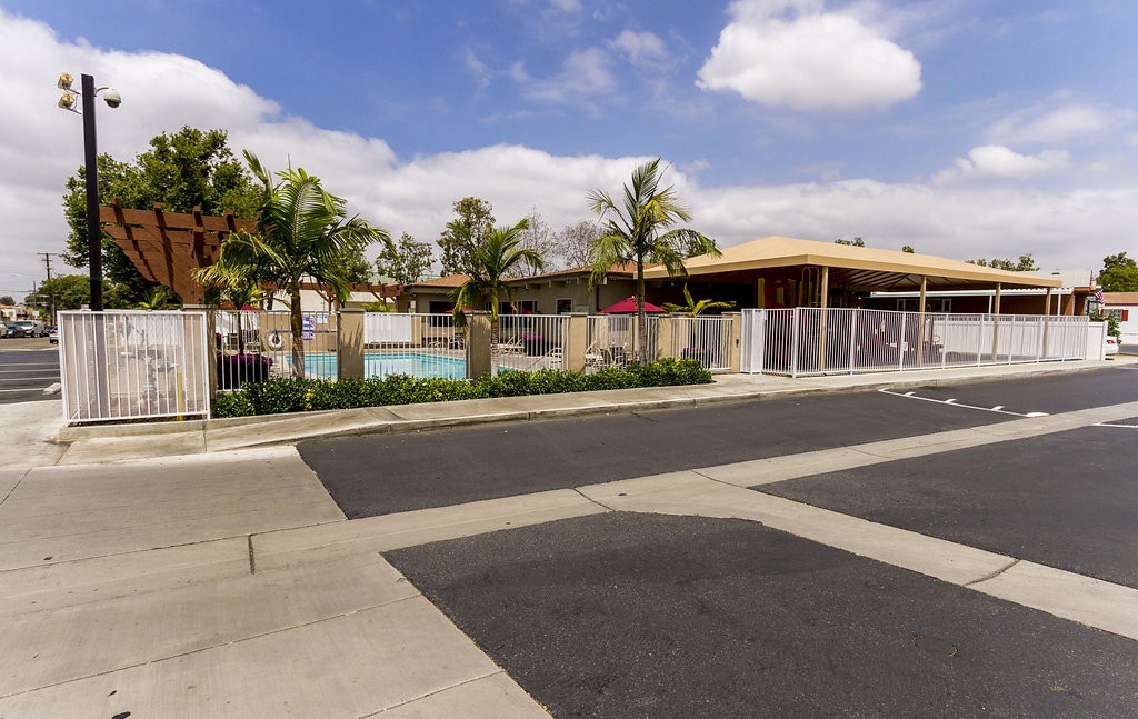 Parking lot view of the outdoor pool and community center. Enclosed by a white gate fence and decorated with palm trees and well maintained landscape.