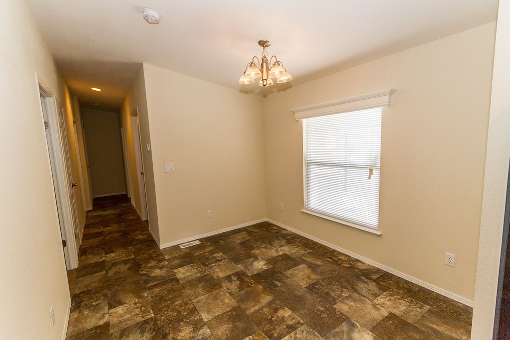 Open space within a home that can be used for a family room, dining room, or extra space. Tile flooring, and cream walls throughout with a small chandelier hanging from the ceiling. Connected is a hallway leading to additional rooms.