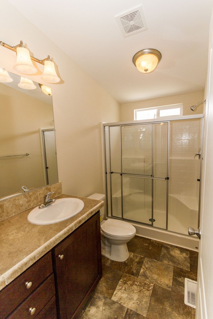 Full bathroom with cream tile-laminate flooring, cream counter tops and single sink with lights above the mirror. Full size walk in shower with single light overhead.