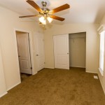 Single bedroom with tan carpet and full size closet. Ceiling fan and light hang overhead in the middle of the room, and single window allows for natural light throughout.