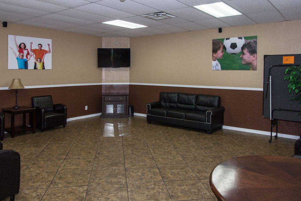 All tile floor in game room at Sierra Estates with flat screen on the wall. Leather couch and chairs and a folded up ping pong table