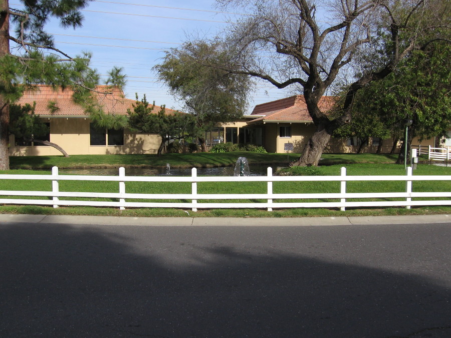 Beautiful view of the community center and pond, sitting behind a white picket fence and surrounded by well-maintained green grass and trees.
