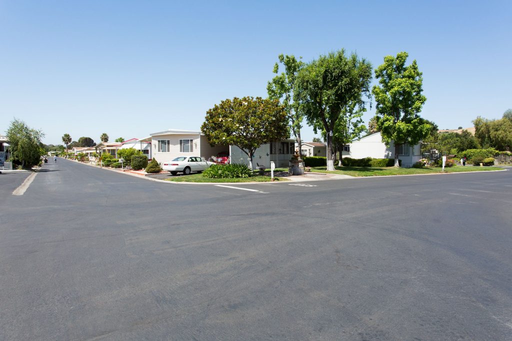 Wide corner street view within the senior community. Beautiful greenery line the streets with multiple manufactured homes.