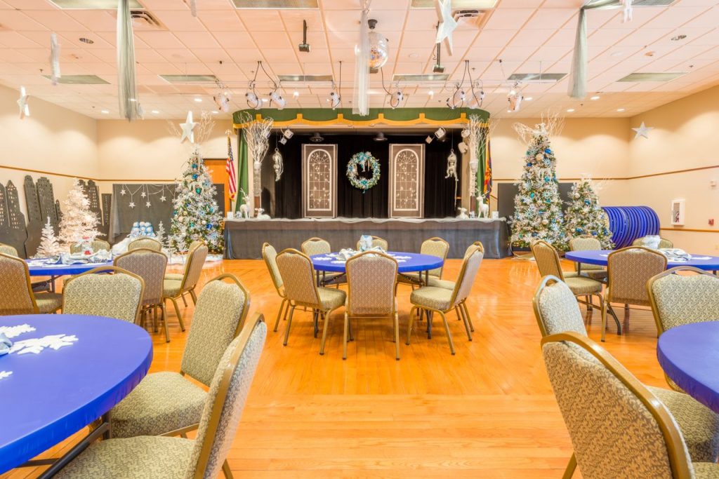 Ballroom decorated for a winder wonderland with 5 Christmas trees decorated in white, blue and green. Round tables decorated with blue tablecloth and white snowflakes and 8 chairs. A stage is decorated in Christmas theme with a blue, green white wreath. White stars also hang from the ceiling.