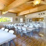 Open community center with high vaulted, wood ceilings and beautiful wood flooring. Spacious area for events. Long fold out tables and chairs make up majority of the space. Windows and ceiling fans with lights create a bright space.