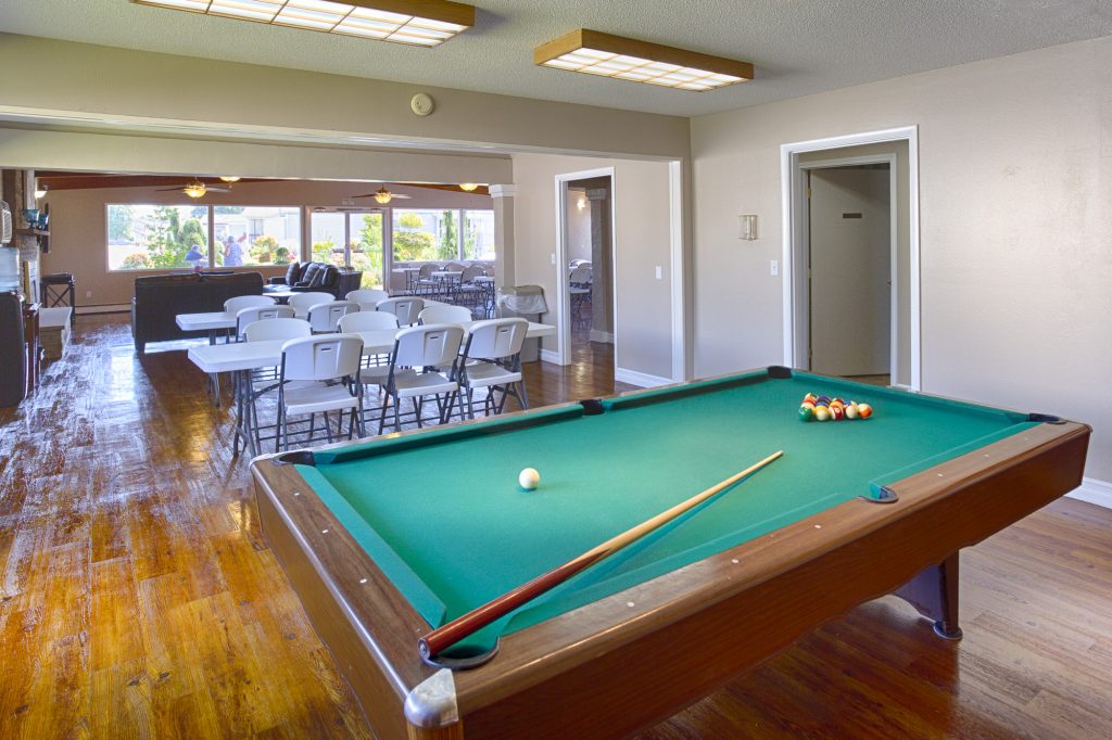 Community center equipped with a pool table and a variety of seating. Walls are lined with windows providing natural light.